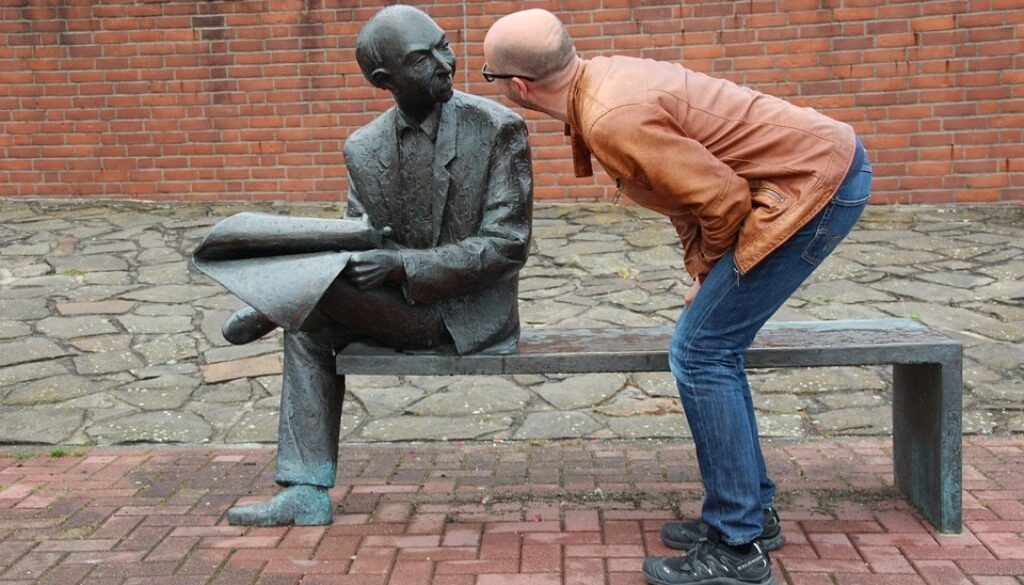 uomo che parla con statua che raffigura un uomo che legge un giornale seduto su una panchina