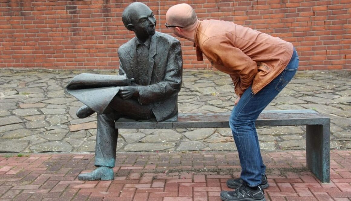 uomo che parla con statua che raffigura un uomo che legge un giornale seduto su una panchina